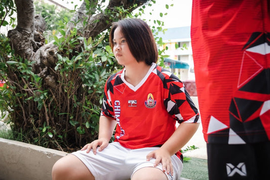Young person in a red sports jersey sits outdoors.